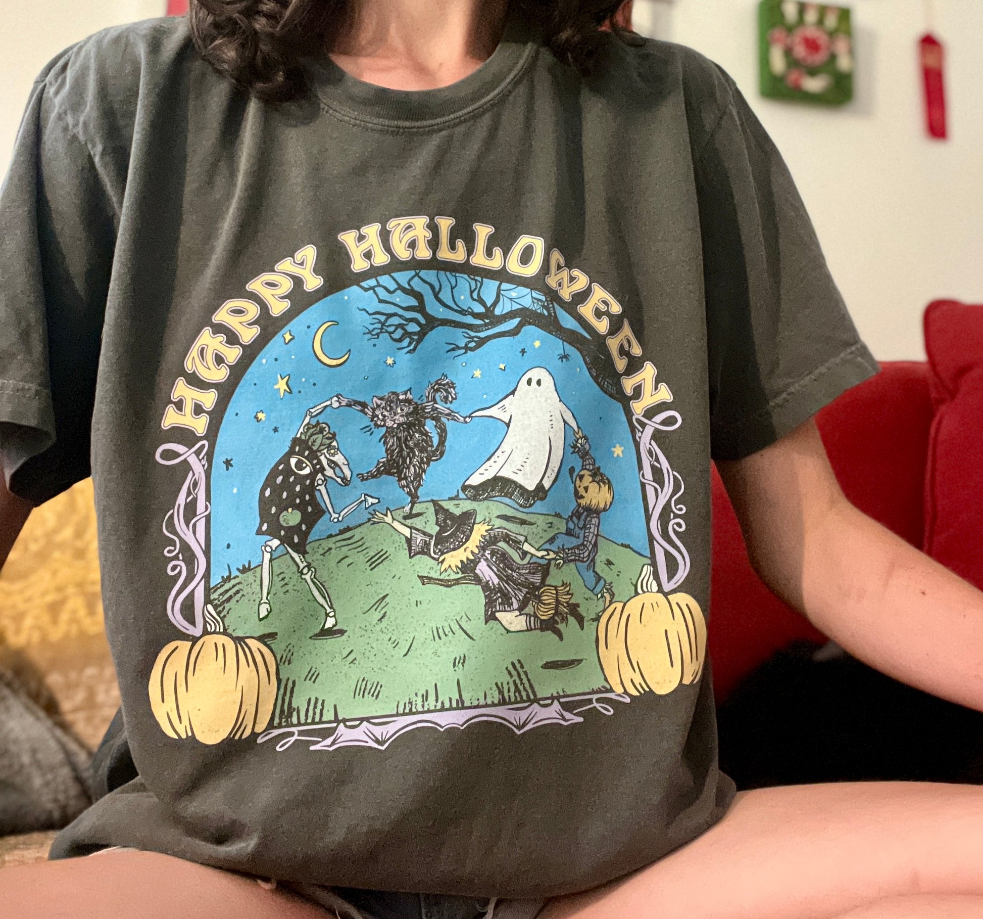 Girl in grey happy Halloween t-shirt. The illustration is cute and spooky and inspired by henri Matisse. It has a skeleton, scare crow, witch, cat, and ghost in a dance circle with tree branches and pumpkins as a border. Says happy Halloween on the top. Cotton t-shirt, comfort colors. Asheville artist. 