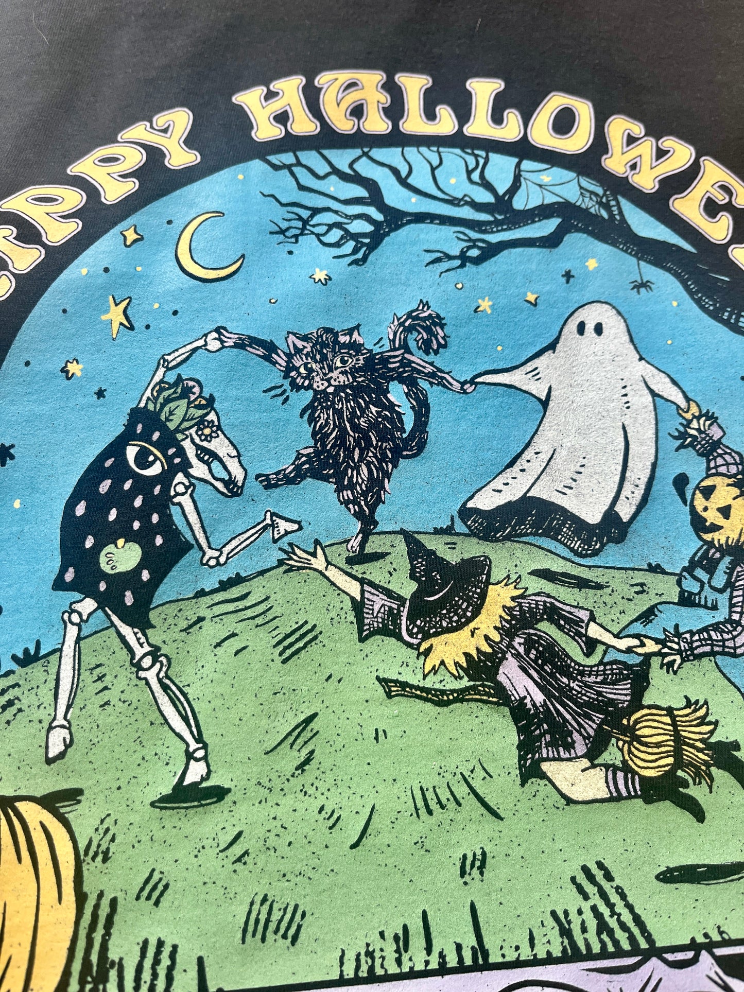 Happy Halloween black t-shirt closeup view. With spooky characters dancing in a circle, inspired by Henri Matisse The Dance painting. Ghost, black cat, witch, pumpkin headed scare crow, and an animal skeleton having a good Halloween time. With pumpkin border and text that says happy Halloween. Asheville artist. 