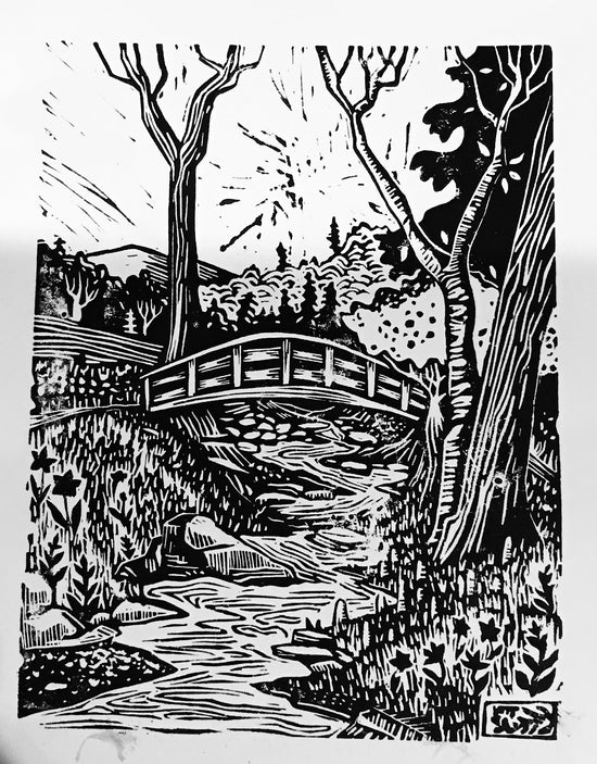 A colorful linocut print depicting a spring landscape with a bridge, trees, and a stream, displayed on a table with a floral patterned background.