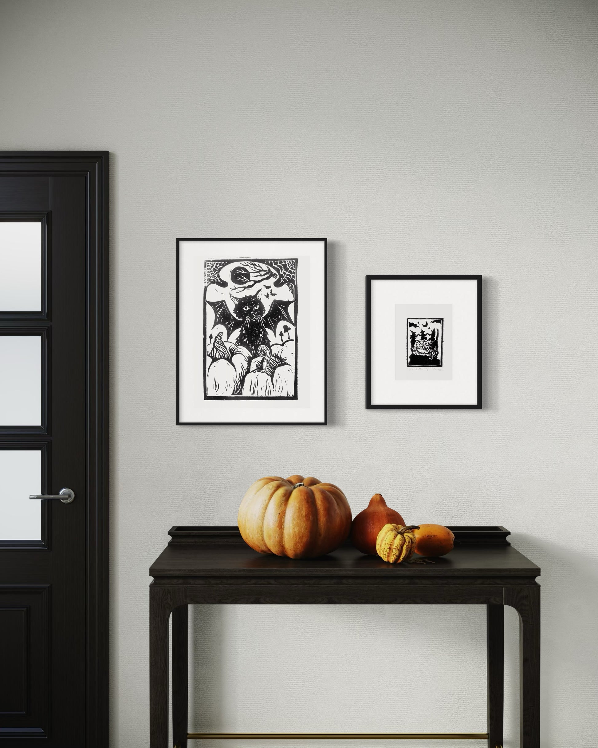 linocut print of a black cat with bat wings surrounded by pumpkins and a linocut of witches dancing around a pumpkin, by asheville artist erika busse framed 